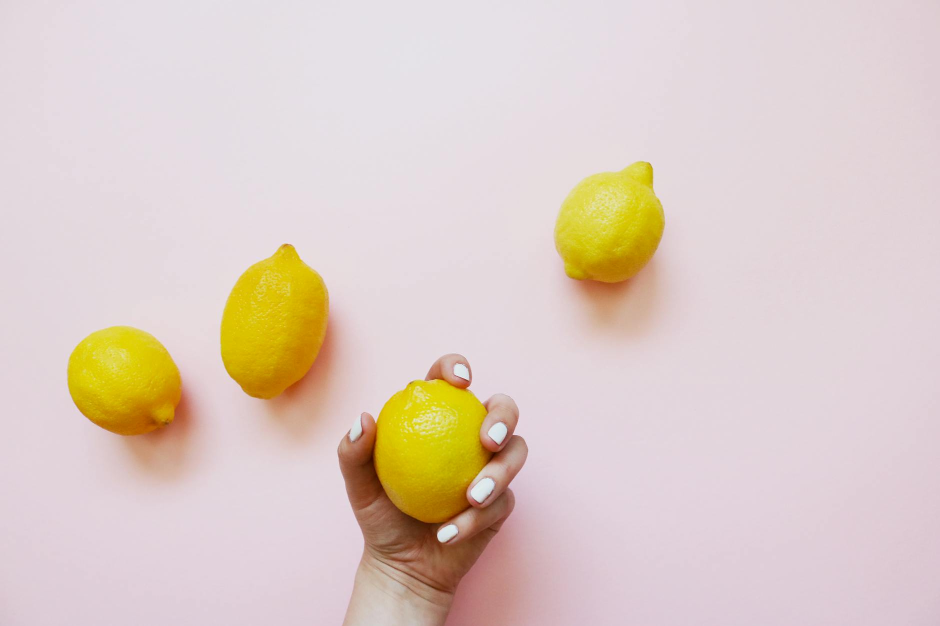 A hand with white nails holding a fresh lemon on a soft pink background, symbolizing pleasure and self-care